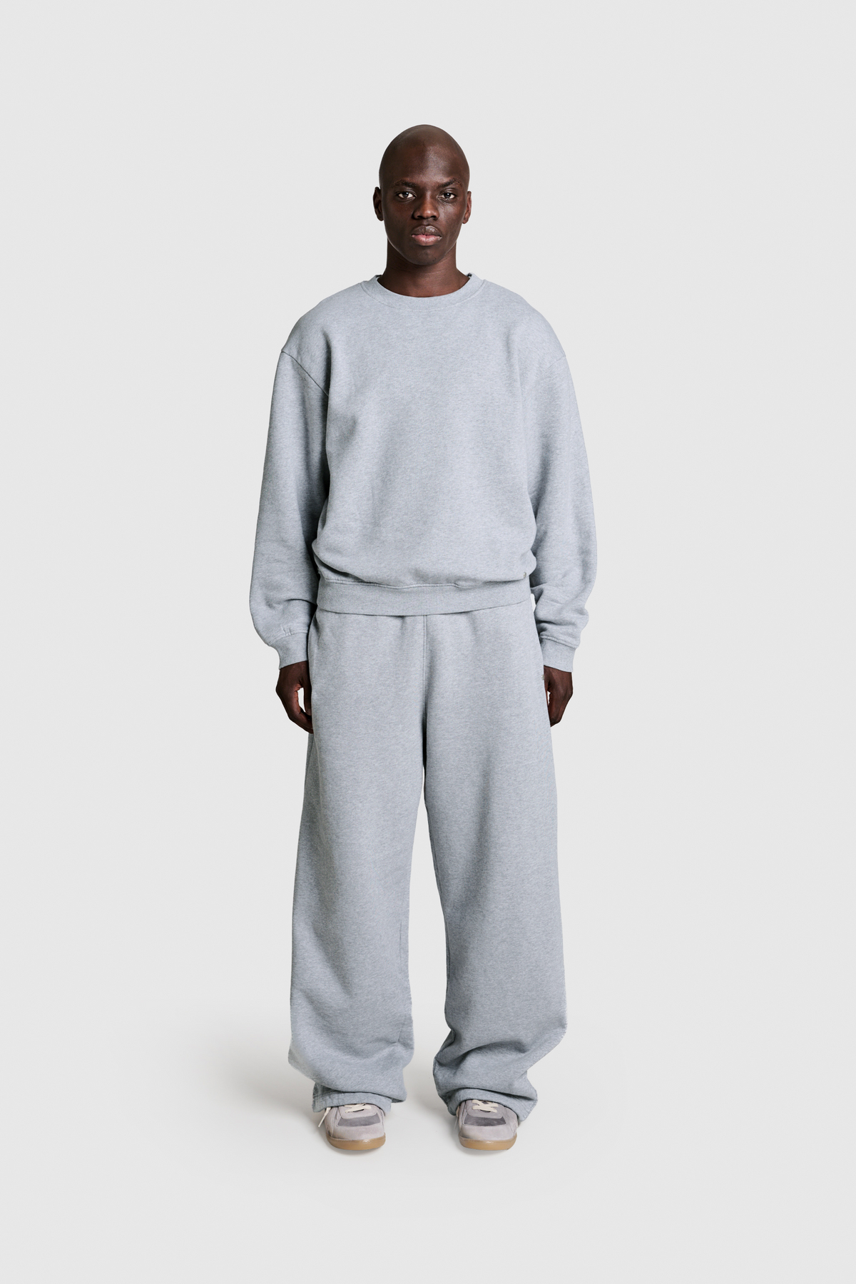 A person stands facing forward against a plain white background, wearing the PESOCLO Raws 2-in-1 Jogger—a loose-fitting gray sweatshirt and matching sweatpants ensemble crafted from 100% Baumwolle with a subtle vintage wash—paired with light-colored sneakers.