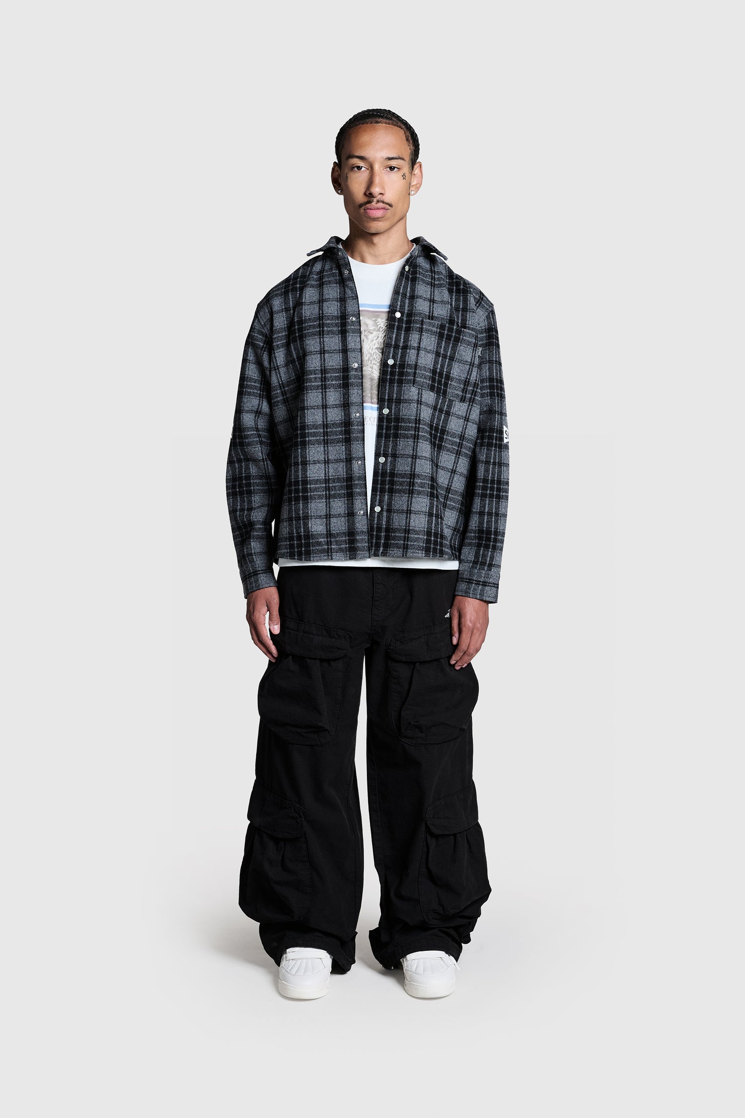A man stands facing forward, wearing the PESOCLO Boulevard Flannel—a gray plaid shirt crafted from Wolle with a Regular Fit and a practical Brusttasche—layered over a white t-shirt, paired with black baggy cargo pants and white sneakers, against a plain light gray background.