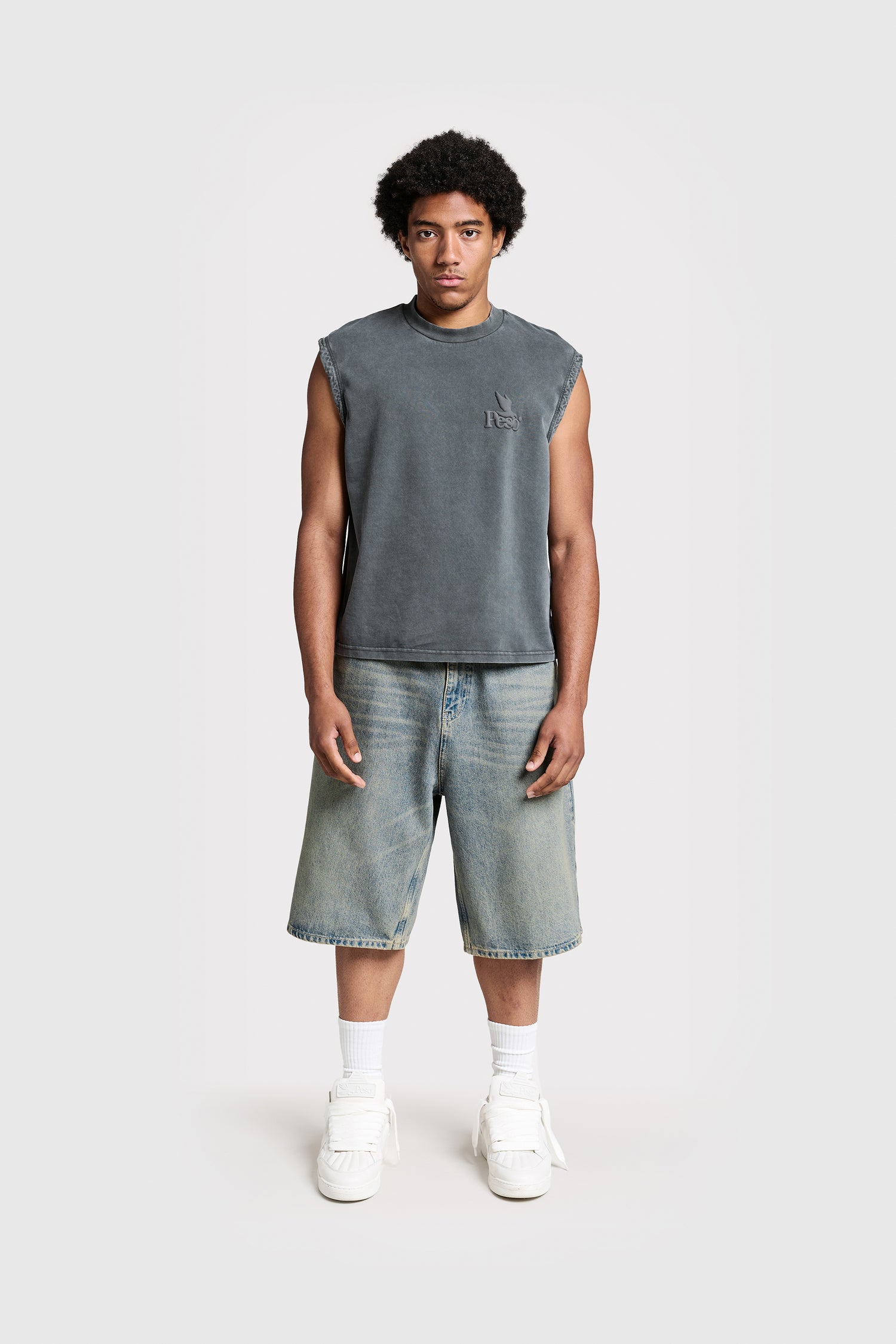A man stands facing forward against a plain light background, wearing a relaxed fit sleeveless gray shirt, PESOCLO Logo Raws Denim Shorts, white socks, and white sneakers.