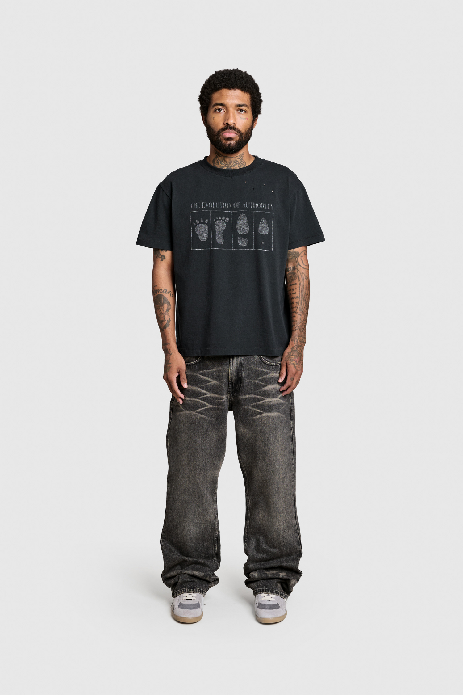 A man with a beard and tattoos stands facing forward against a plain white background, wearing the PESOCLO Evolution Tee—a black graphic T-shirt made from 100% Baumwolle—paired with loose-fitting faded jeans with a vintage wash and gray sneakers.