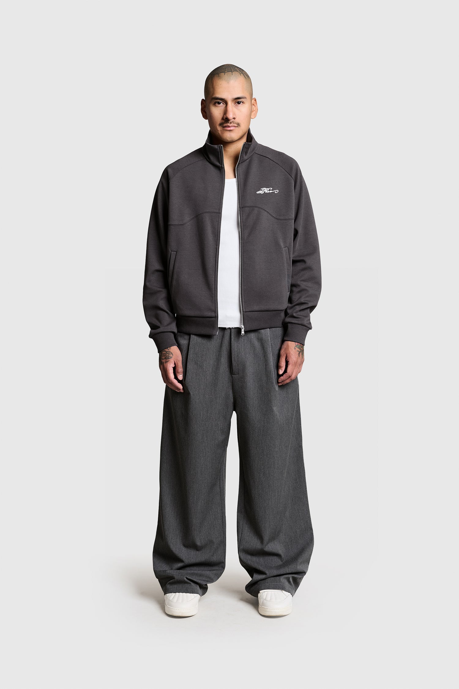 A person with short hair stands facing forward against a plain white background, wearing the PESOCLO Casa Trackjacket—a gray zip-up jacket crafted from a comfortable Polyester Baumwolle blend—paired with a white shirt, wide gray Regular Fit pants, and white sneakers.