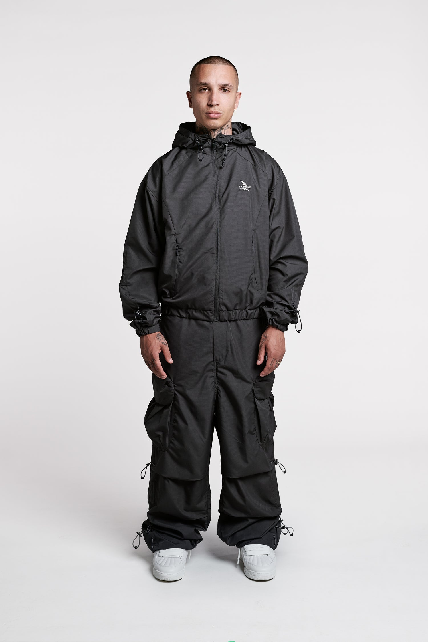 A person stands facing forward, wearing a black hooded polyester jacket and PESOCLO Parachute Pants in black, which feature an elastic drawstring for a comfortable, loose fit, paired with white sneakers against a plain white background.