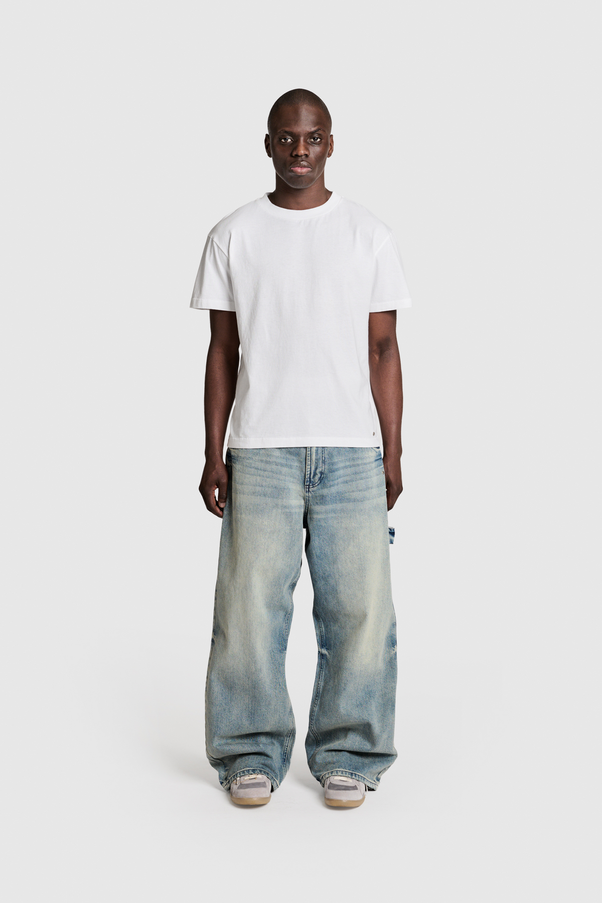 A person stands against a plain white background, wearing the PESOCLO Raws Tee made from 100% Baumwolle, paired with light blue oversized jeans featuring subtle vintage pilling and sneakers.