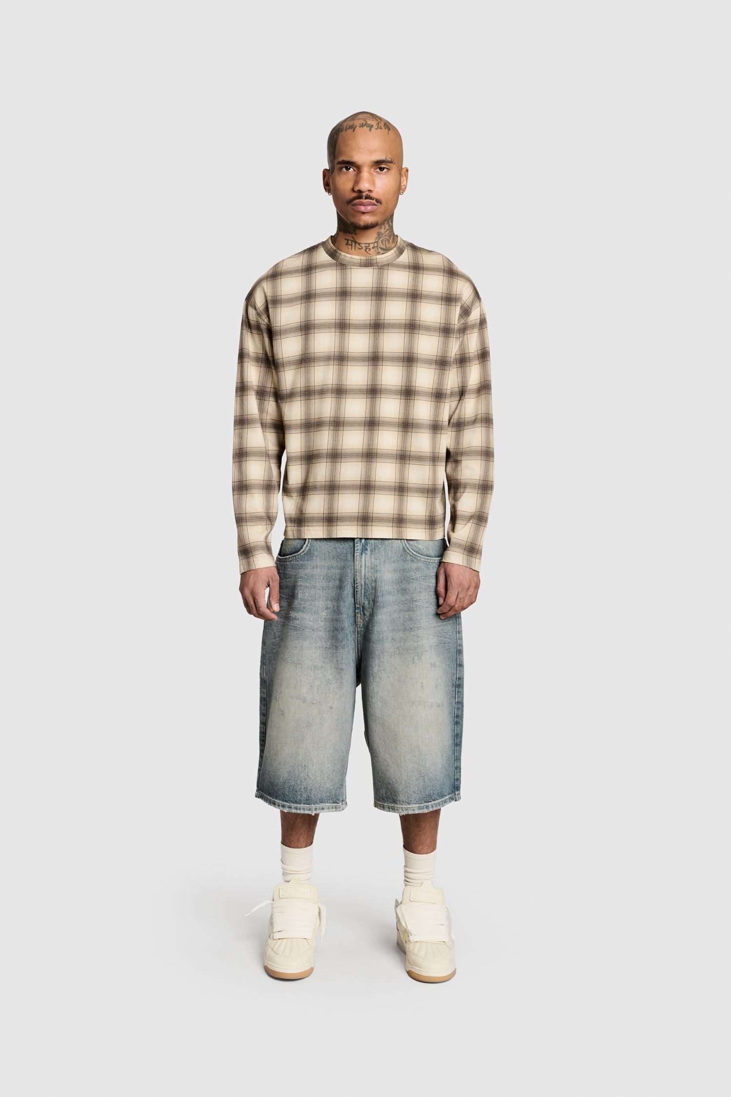 A person stands facing forward against a plain light gray background, wearing the PESOCLO Plaid Longsleeve in beige and brown, paired with oversized faded denim shorts crafted from 100% Baumwolle in a vintage wash and white high-top sneakers.