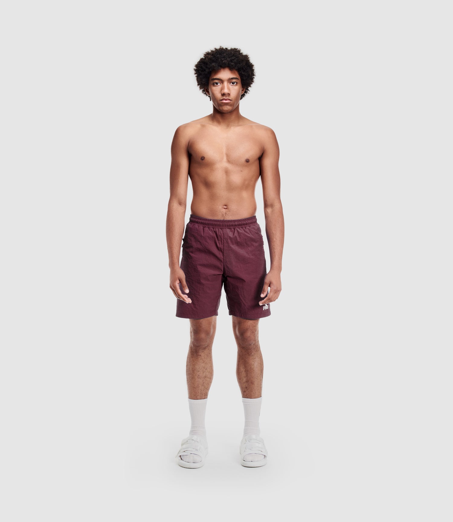 A young man with curly hair stands facing forward, wearing maroon PESOCLO Eagle Swimshorts with an elastic waistband, white socks, and white sandals against a plain white background.