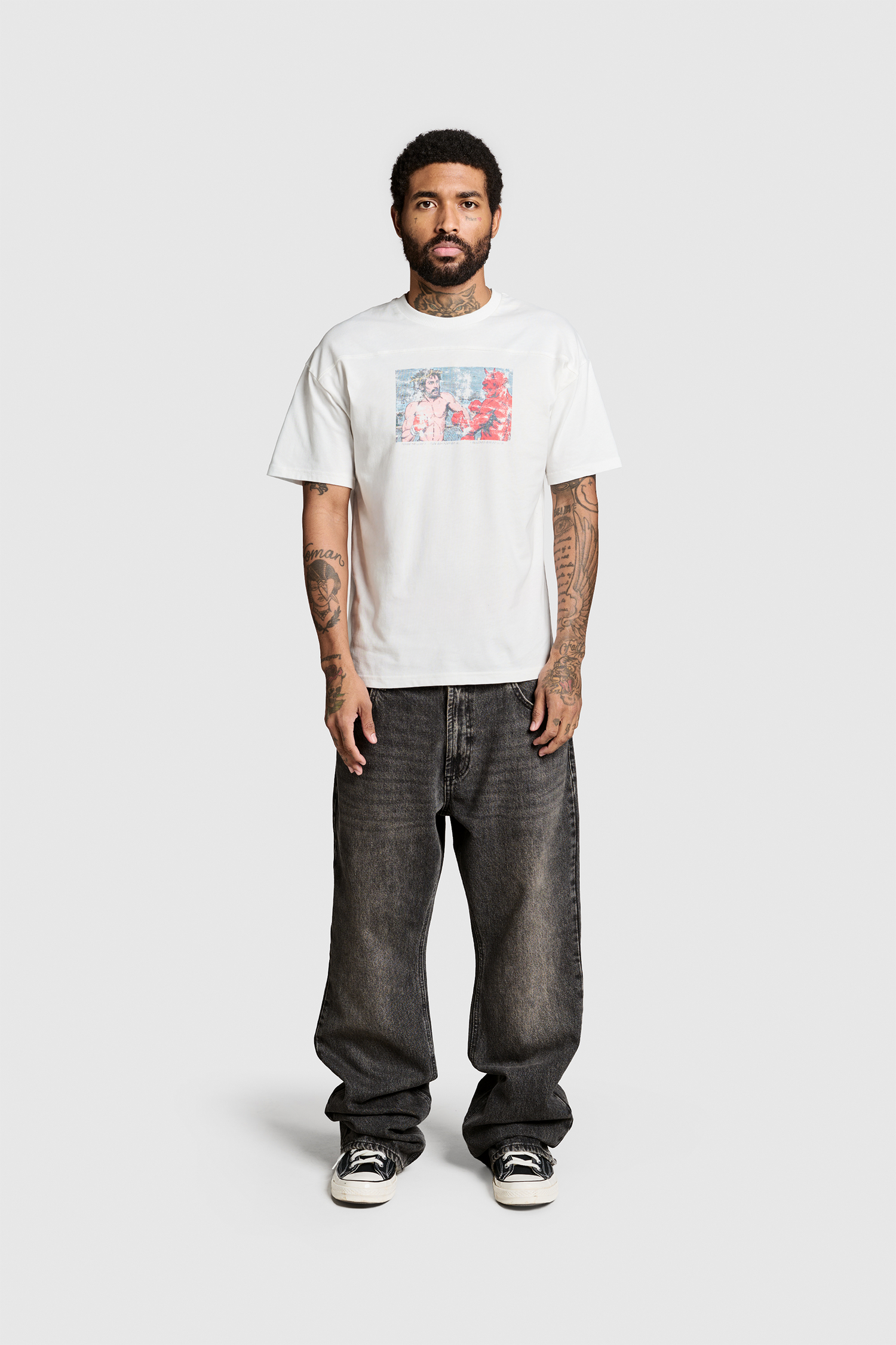 A man with short curly hair and a beard wears the JESUS WINS TEE by PESOCLO—a white graphic T-shirt featuring a striking Frontprint, crafted from 100% Baumwolle—paired with loose-fitting dark jeans in a Regular Fit and black-and-white sneakers, standing against a plain light gray background.
