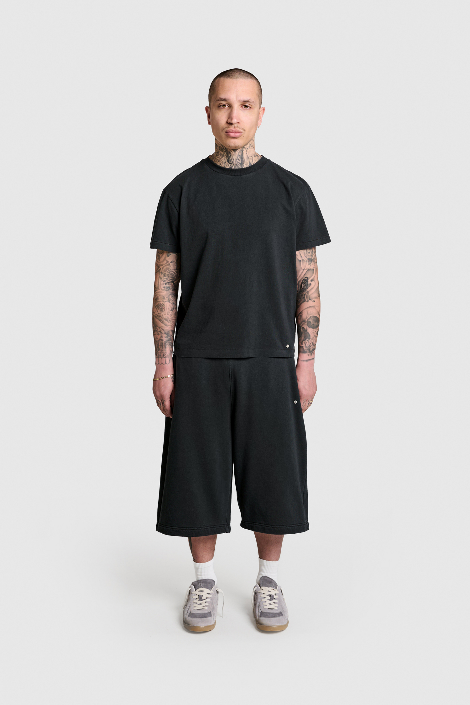 A person with tattoos stands facing forward against a plain light gray background, wearing the PESOCLO Raws Tee—a black t-shirt adorned with a subtle metal logo patch—paired with black knee-length shorts, white socks, and gray sneakers. The outfit is crafted from 100% Baumwolle for comfort and features a regular fit for effortless style.