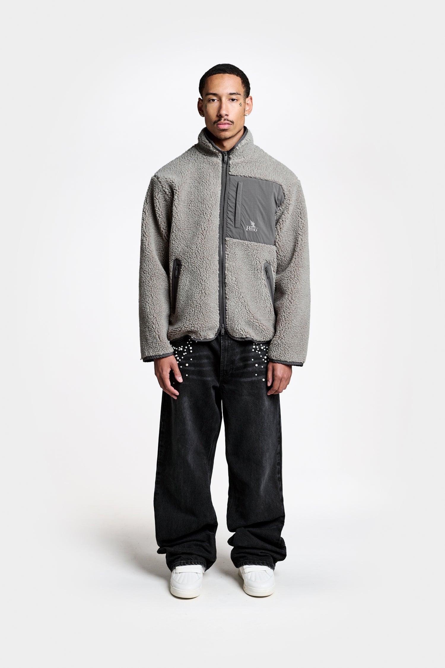 A man stands facing forward against a plain white background, wearing the PESOCLO Sherpa Fleece—a light gray zip-up jacket with a tonal embroidered gray chest pocket—paired with baggy black jeans featuring white detailing and classic white sneakers.