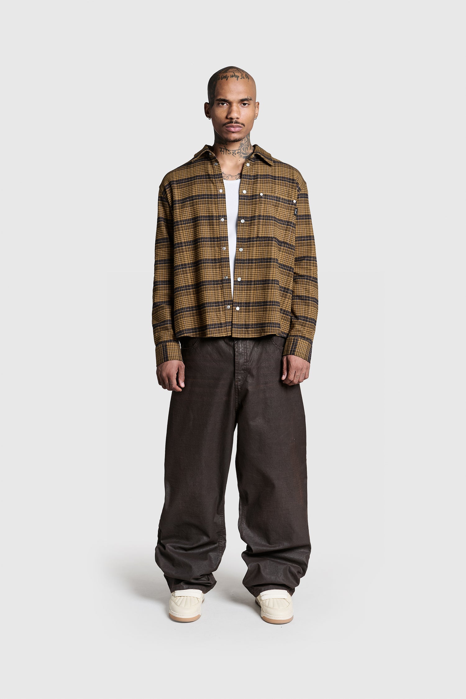 A person stands facing forward against a plain light gray background, wearing the PESOCLO Boulevard Flannel—a brown plaid button-up shirt crafted from 100% Baumwolle—layered over a white top, paired with loose dark brown Regular Fit pants and light-colored sneakers. A subtle Flaglabel detail completes the look.