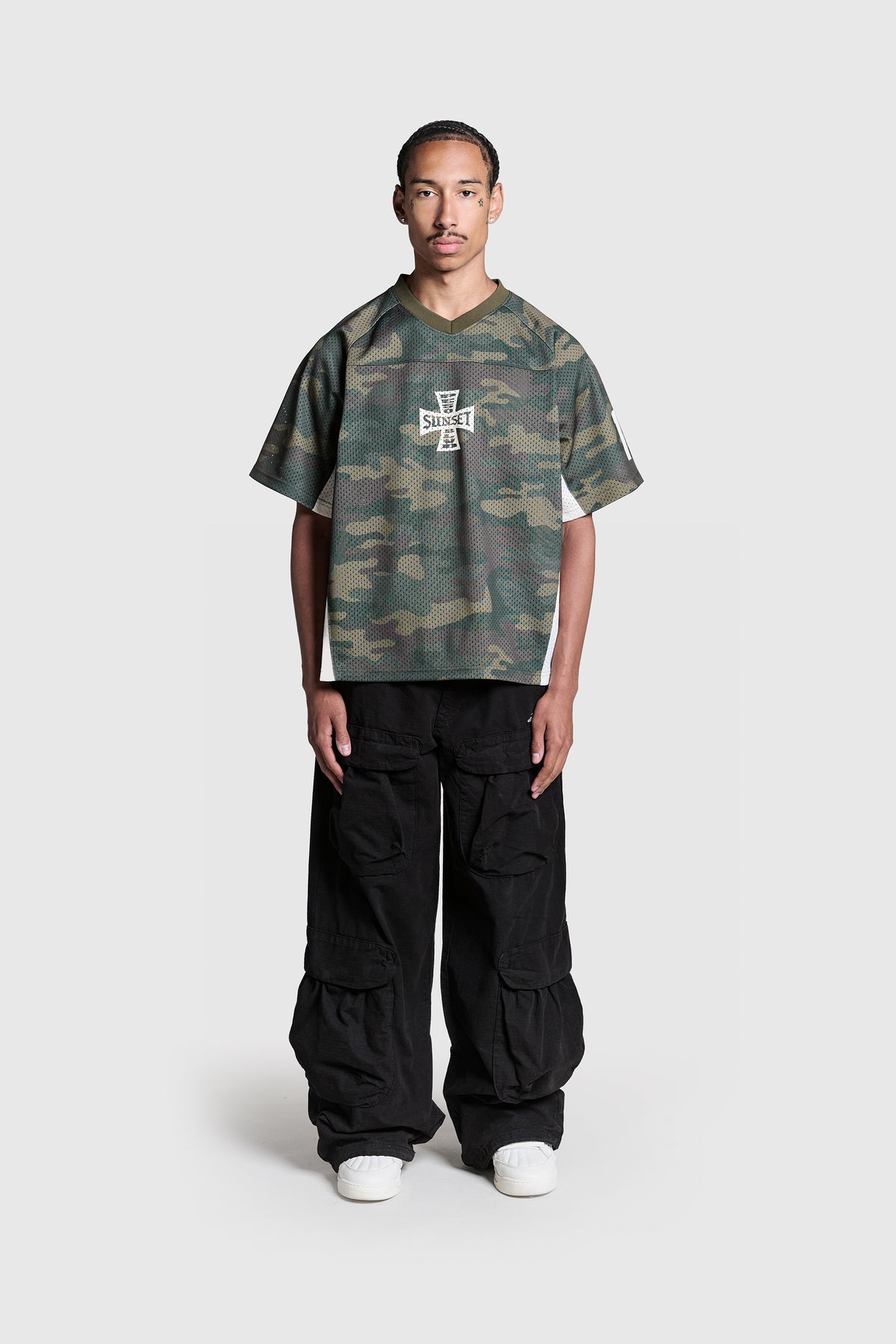 A man stands facing forward against a plain light gray background, wearing the PESOCLO Rose Bowl Jersey—a loose-fit, camo print short-sleeve shirt made from 100% polyester for comfort and durability—paired with black cargo pants and white sneakers.
