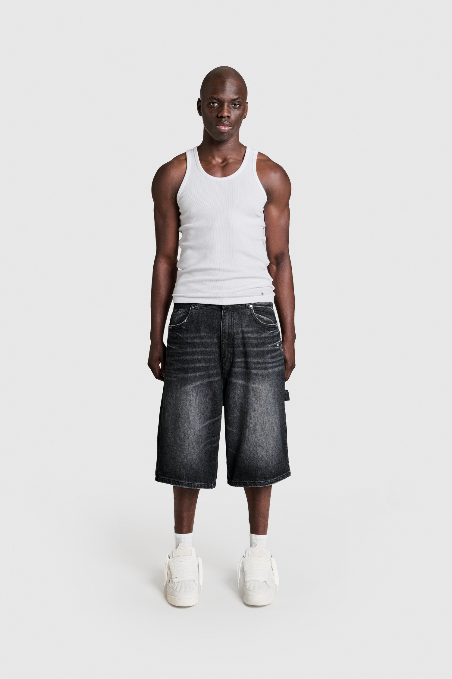 A man stands facing forward against a plain white background, wearing a white tank top and PESOCLO Raws Denim Shorts in a loose fit dark wash with a subtle metal logo patch, paired with white sneakers.