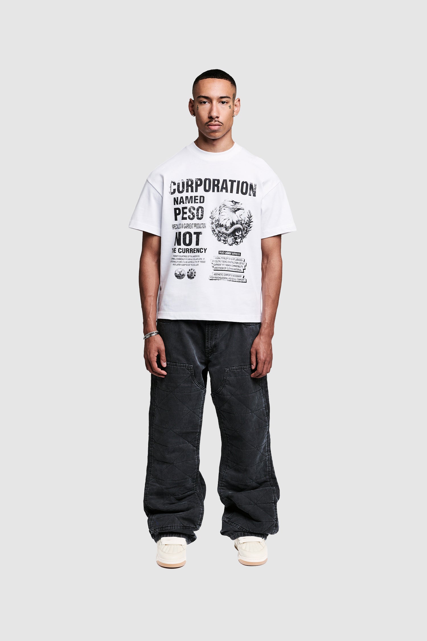 A man stands against a plain gray background, wearing a white graphic T-shirt with black text and images, PESOCLO Carpenter Pants with a vintage wash and baggy fit, and white sneakers. He is facing forward with a neutral expression.
