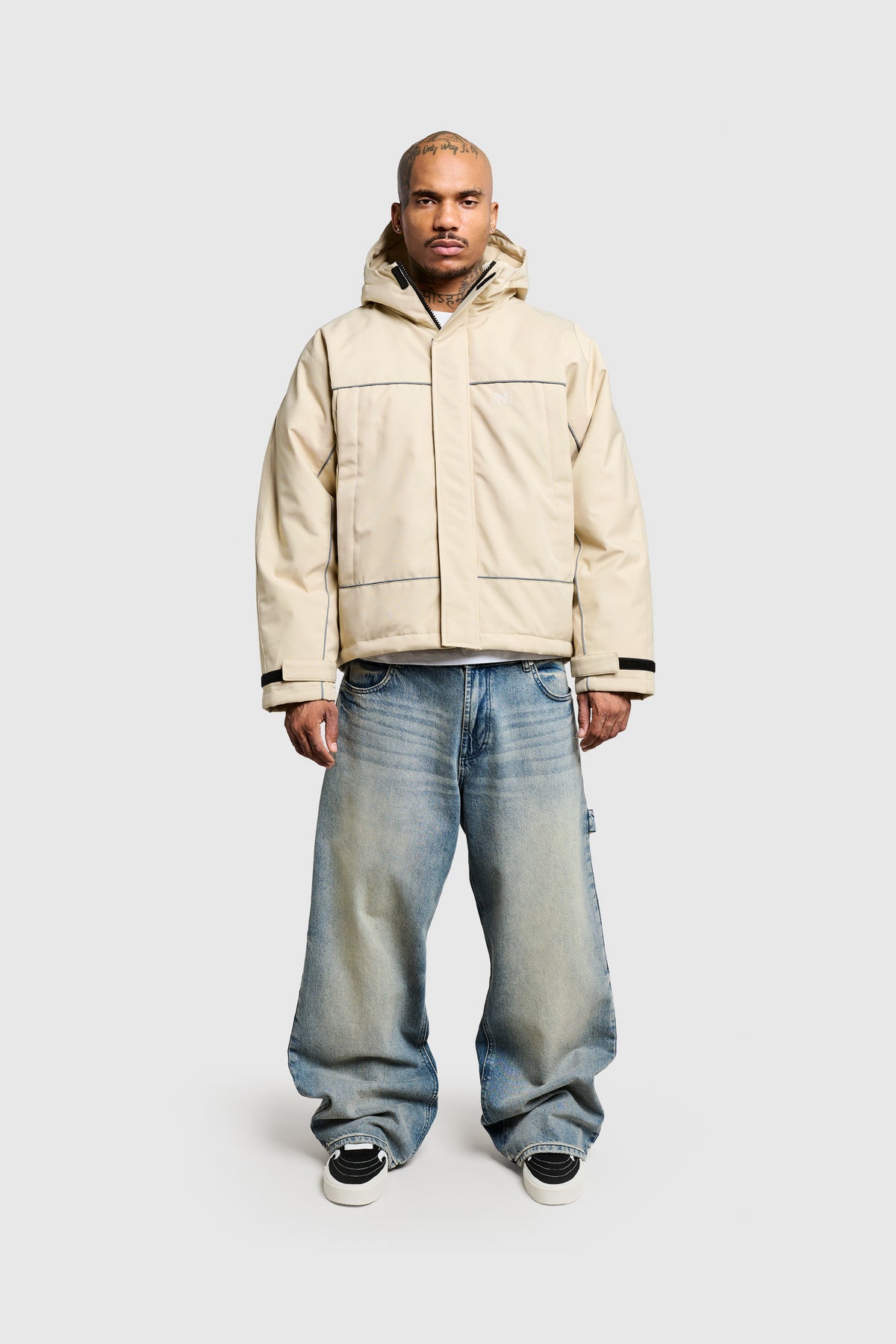 A man with head tattoos stands facing forward, wearing a cream PESOCLO Rainjacket in a regular fit with dark piping and a subtle faux-fur collar. He pairs it with baggy light-wash denim jeans and black sneakers against a plain light gray background.
