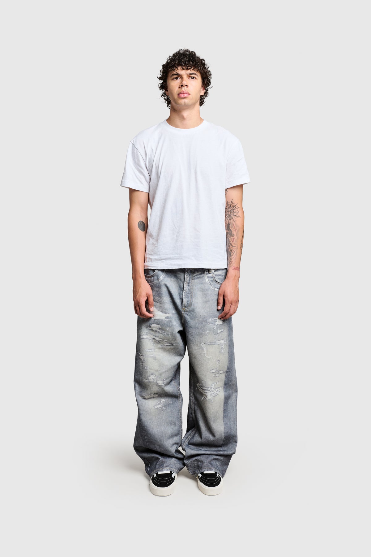 A young man with curly hair stands against a plain background, wearing a plain white t-shirt, black skate shoes, and baggy fit PESOCLO Printed Denim jeans made from 100% Baumwolle. He has tattoos on both arms.