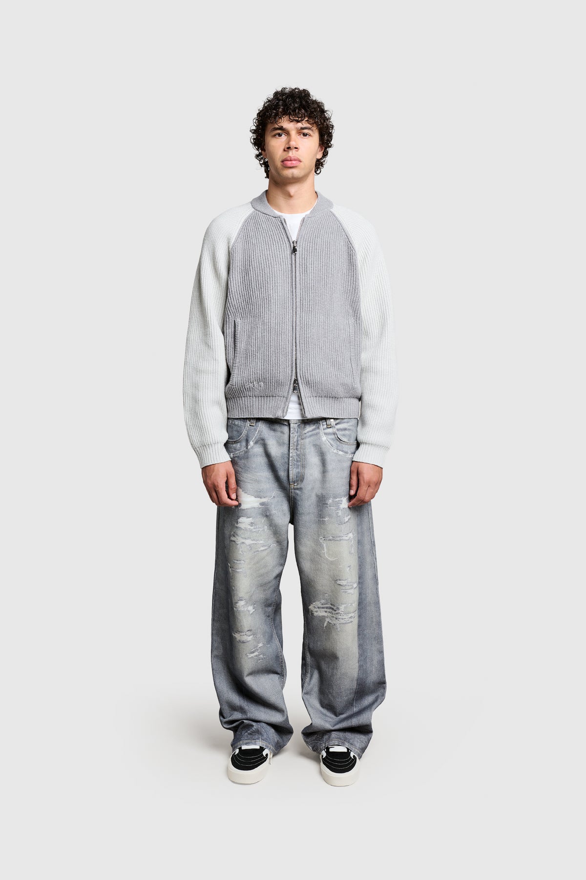 A person with curly hair stands facing forward, wearing the PESOCLO Raglan Varsity light gray zip-up jacket featuring a Zweiwege Reißverschluss and handgesticktes Logo, paired with loose-fitting distressed jeans in a Regular Fit and black sneakers against a plain, light background.