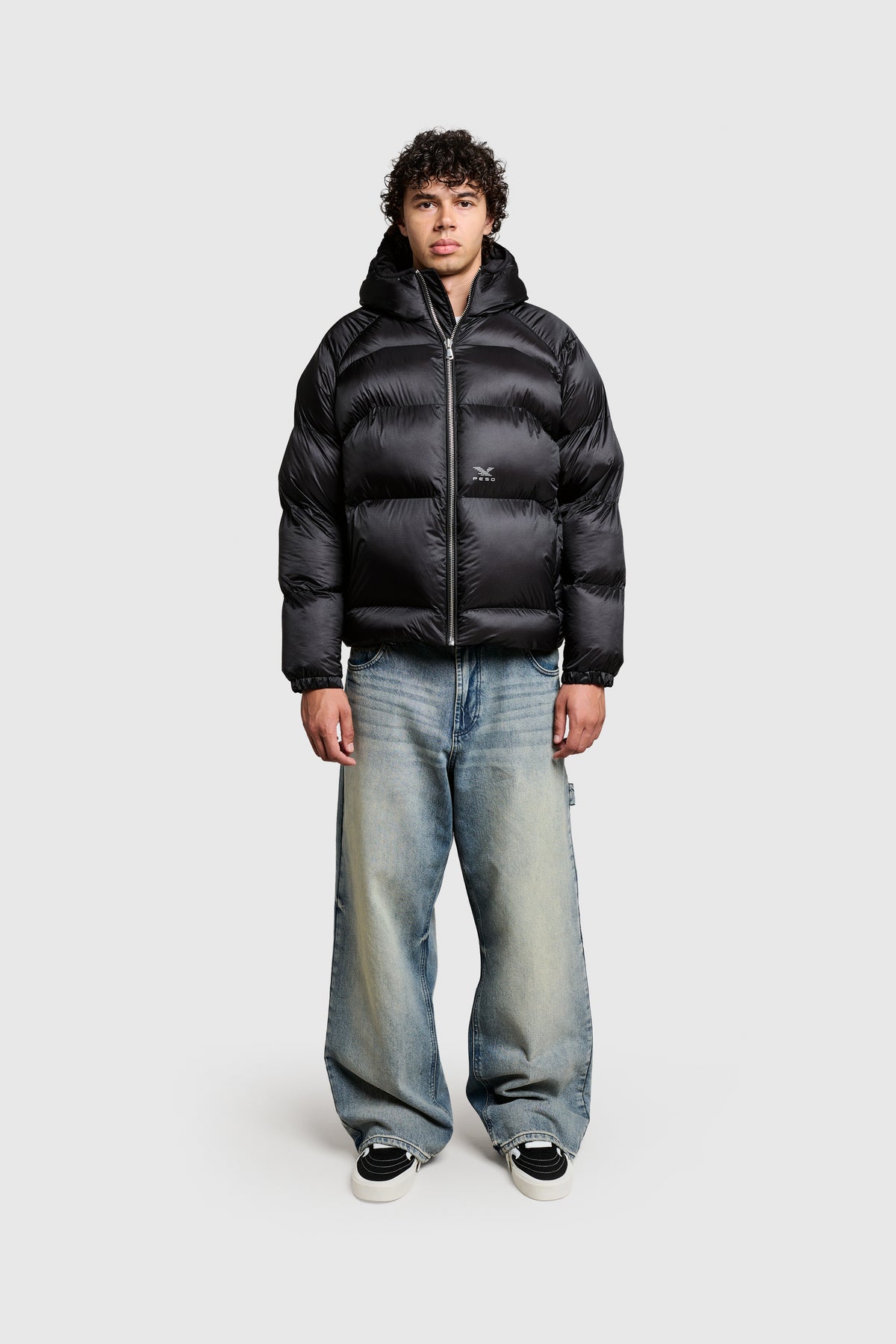 A person wearing the PESOCLO Transclucent Puffer—a black polyamid jacket in a regular fit with a subtle front print—pairs it with light blue baggy jeans and black sneakers, standing facing forward against a plain light gray background. This jacket combines style and comfort with its durable construction.