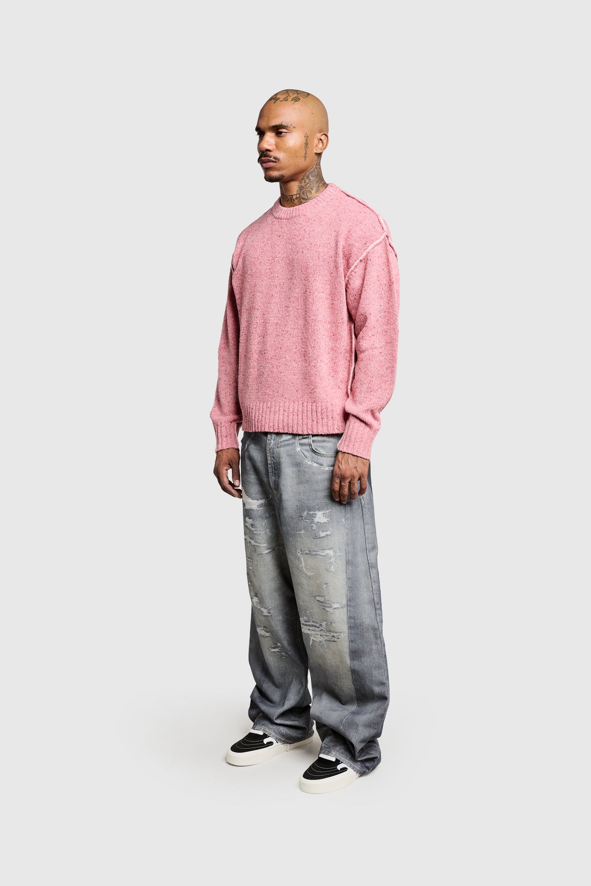 A person stands against a light gray background, wearing PESOCLO’s Raw edge Knit sweater with Optik Rib Bündchen in pink, paired with baggy distressed gray jeans in a regular fit and black-and-white sneakers. The person has short hair and visible tattoos on the neck and head.