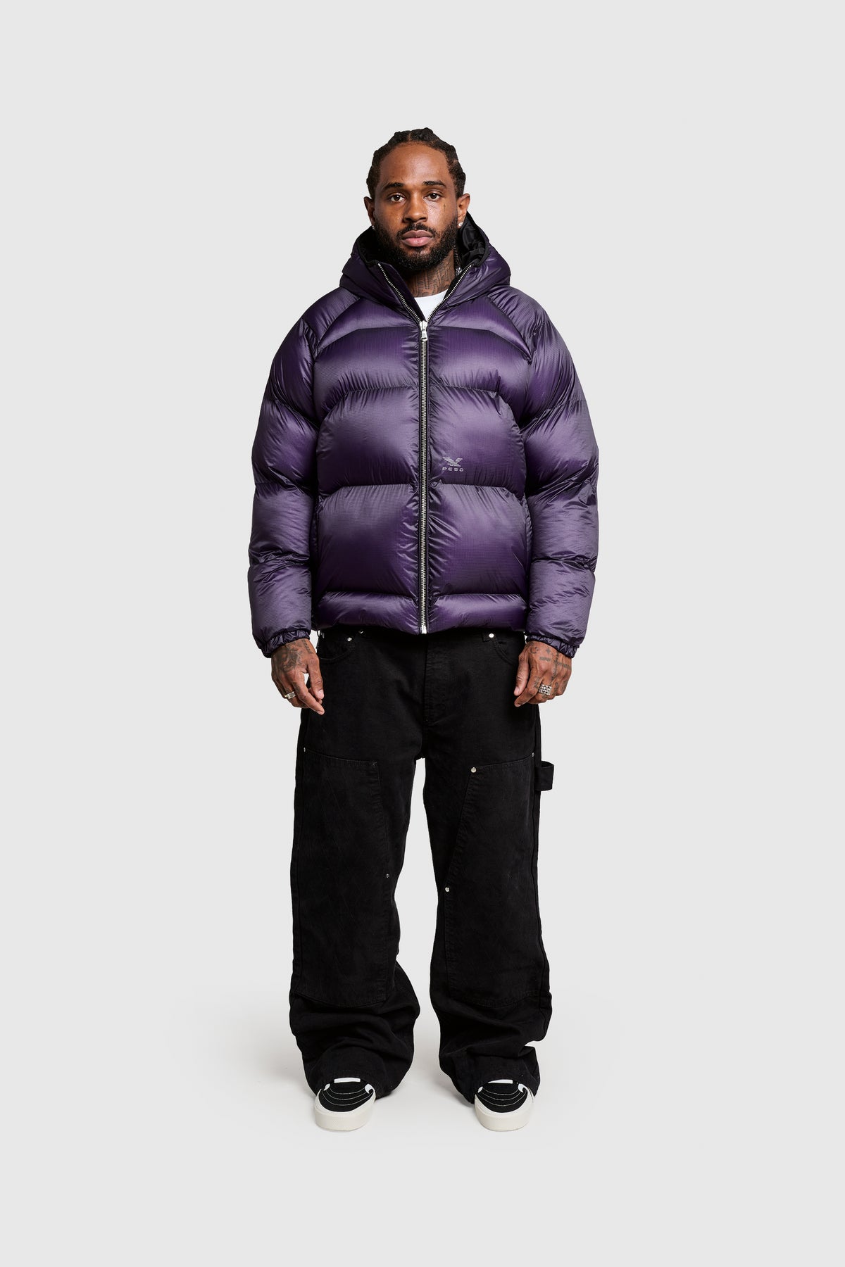 A man stands facing forward against a plain white background, wearing the PESOCLO Transclucent Puffer—a shiny purple polyamide puffer jacket featuring a front print—paired with black cargo pants and black sneakers with white soles.
