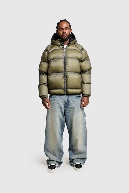 A man stands facing forward, wearing a shiny olive green Transclucent Puffer from PESOCLO with a hood, baggy light blue jeans, and black-and-white sneakers, against a plain light gray background.