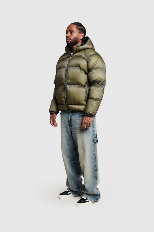 A man stands against a plain white background wearing the PESOCLO Transclucent Puffer in olive green, featuring durable Ripstop-Gewebe, paired with faded loose jeans and black-and-white sneakers.